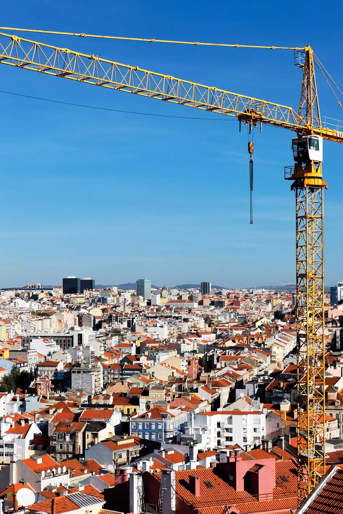 A tall yellow lattice tower crane positioned over a vast city of red-roofed buildings under a clear blue sky.